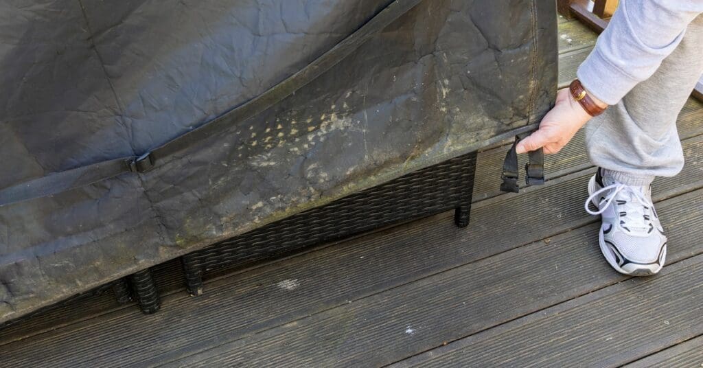 A close-up of a person pulling a black patio cover over a black wicker chair on a dark-colored wooden deck.
