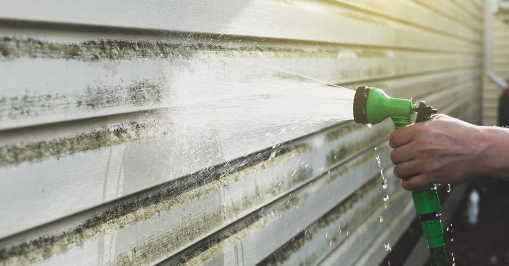 A close-up of someone using a hose with a lime green trigger nozzle to spray beige house siding covered in algae and mold.