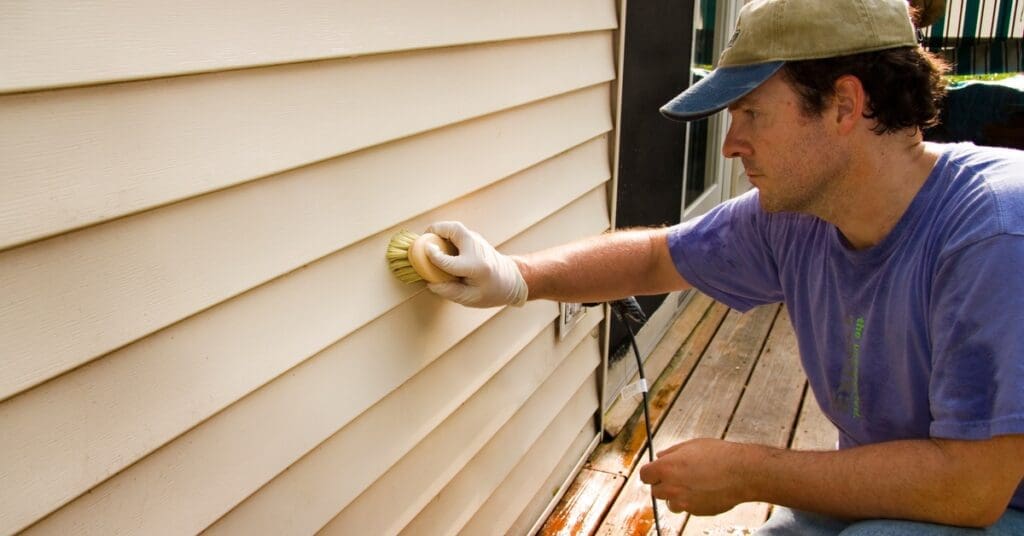 A man in a wet purple T-shirt and denim pants brushing beige vinyl siding near a sliding door with his gloved right hand.