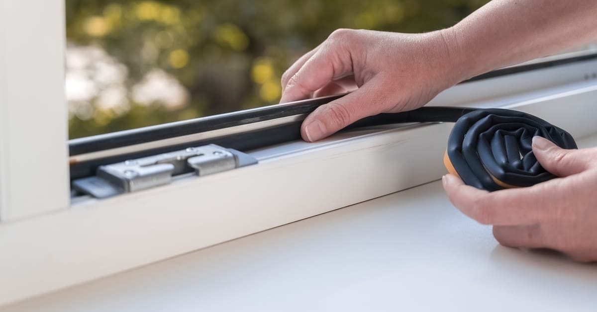 A person presses black sealant into the edge of a white window frame with trees visible through the glass.
