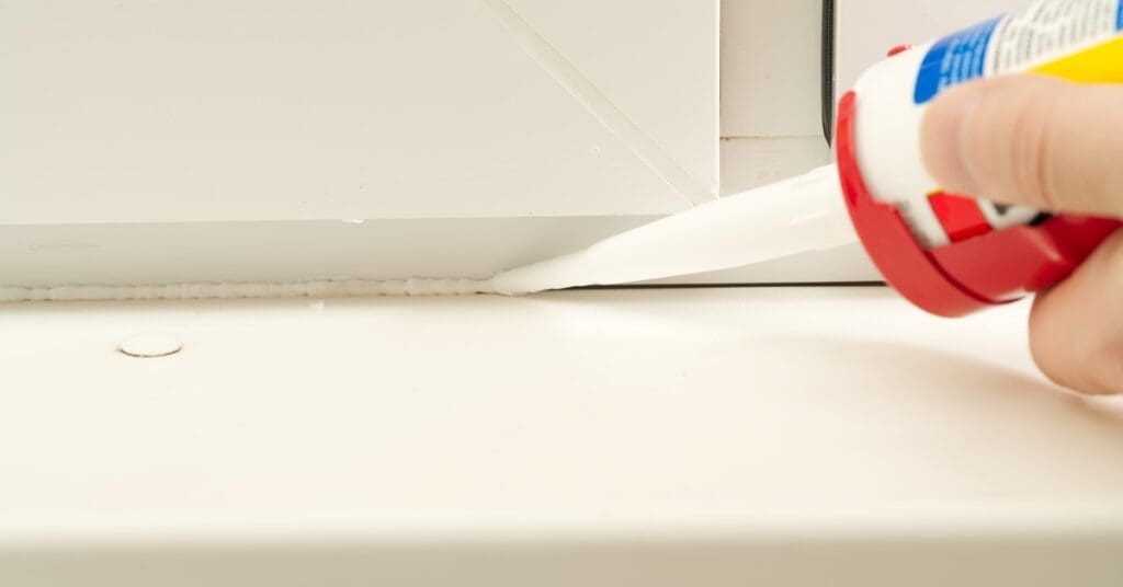 A person applies silicone sealant into a narrow gap between a white window frame and a white window sill.
