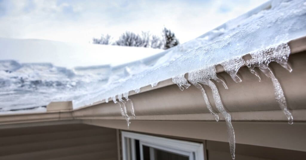 Ice coats taupe box gutters along a snow-covered roof on a house with taupe siding and visible ice at the roof edge.