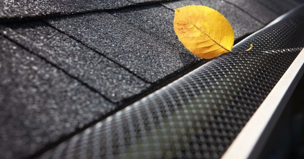 A yellow leaf rests on a plastic gutter cover beneath black asphalt shingles and above a white metal gutter.