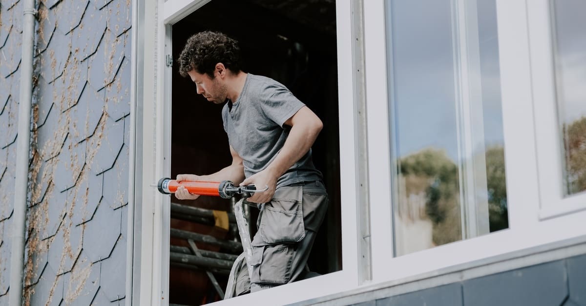 A man standing inside of a home reaches through an open window to caulk the exterior window frame to prevent water damage.