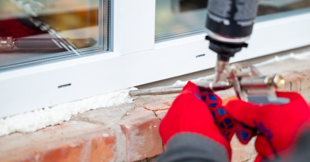 A man wearing red gloves holds a caulk gun as he applies caulk around the exterior side of a home's window.