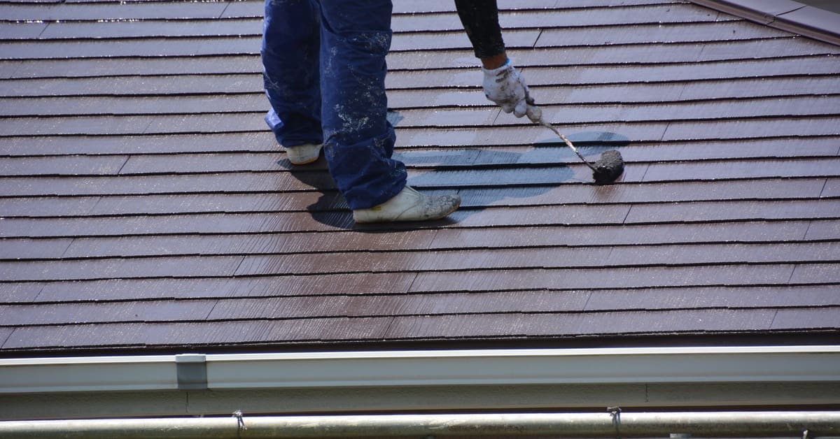A close-up view of the bottom half of a contractor standing on a roof as they bend over to apply a roof coating.