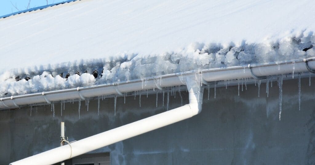 A home in the winter with a roof completely covered in snow. Large chunks of ice accumulate in the gutters.