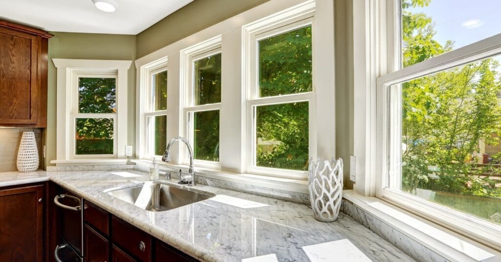 A bright kitchen contains a white marble countertop, dark wood cabinets and large windows with a view of green trees outside.