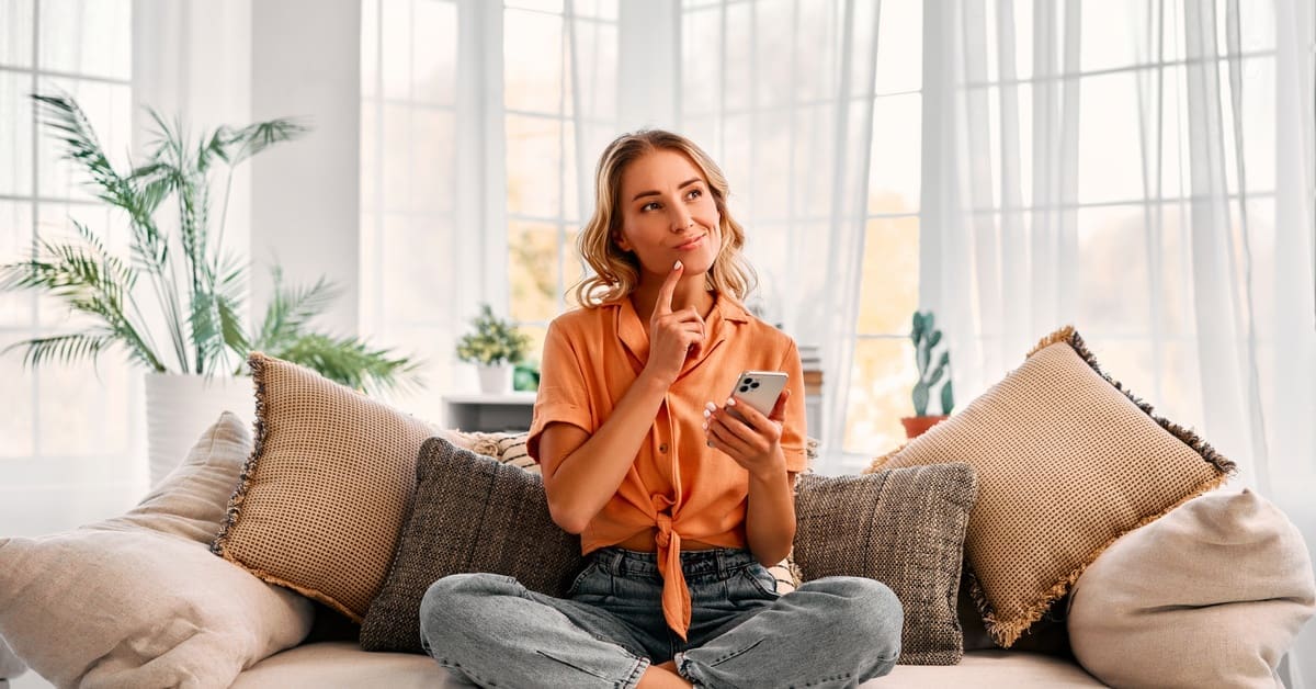 A woman wearing an orange top and blue pants sits on a sofa in her living room, as she touches her chin to think.