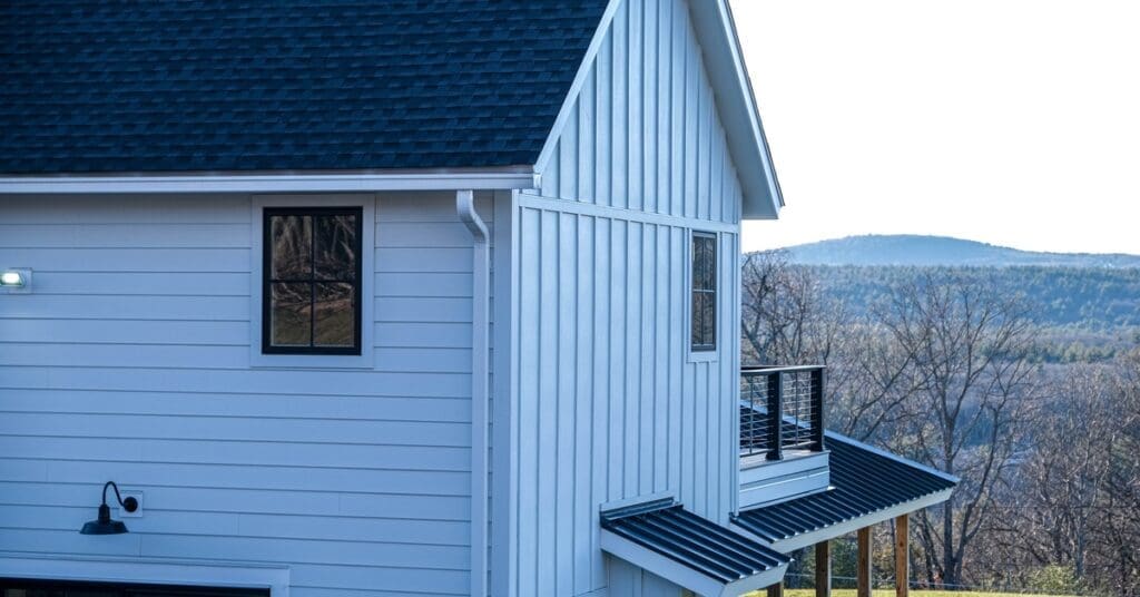 A modern home contains black and white fiber cement siding. There is a forest and rolling hills in the background.