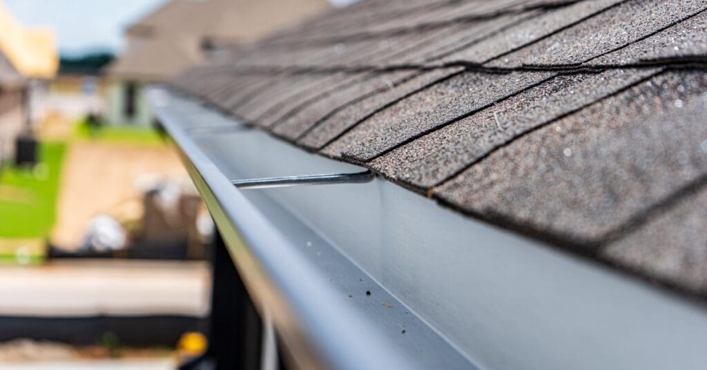 A close-up view of a home containing clean, silver metal gutters and a black, textured asphalt shingle roof.