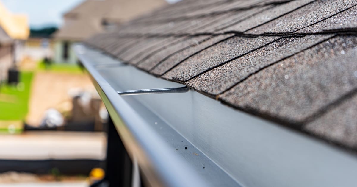 A close-up view of a home containing clean, silver metal gutters and a black, textured asphalt shingle roof.
