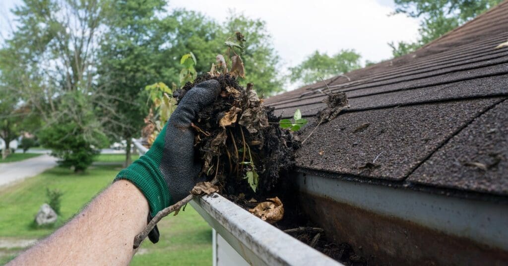 A gloved hand removes wet debris from a metal gutter on a house, with a shingle roof and blurred trees in the background.
