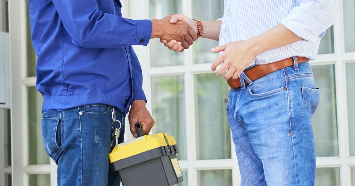 A contractor wearing a blue uniform and holding a tool box shakes hands with a client in front of a glass door.