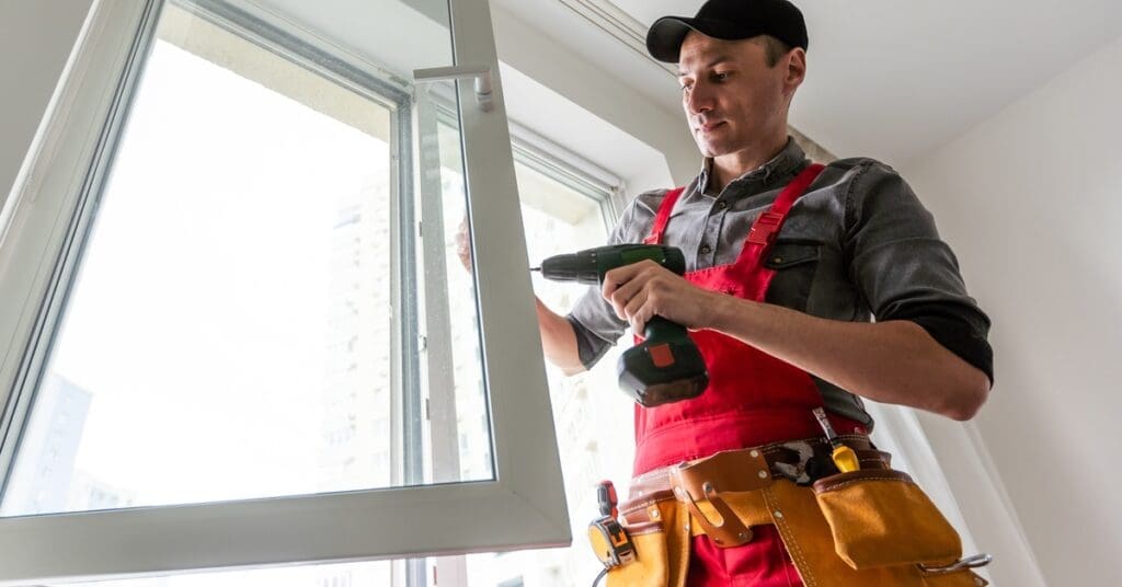 A contractor wearing red overalls uses a cordless drill to install a white-framed window in a brightly lit room.