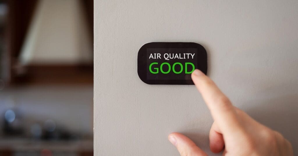A hand points to a black air quality monitor on a light gray wall. The monitor's display reads "AIR QUALITY GOOD"