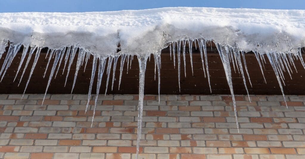 A snow-covered roof has multiple ice dams forming along the gutter. A clear blue sky is visible above the house.