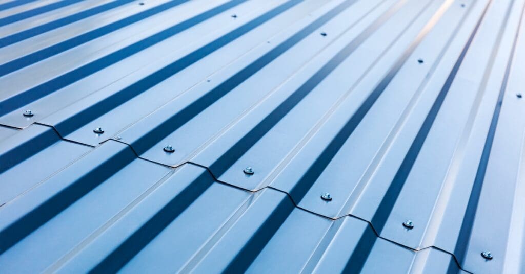 A close-up of a blue metal roof containing visible ridges, valleys, and fasteners reflects light under angled sunlight.