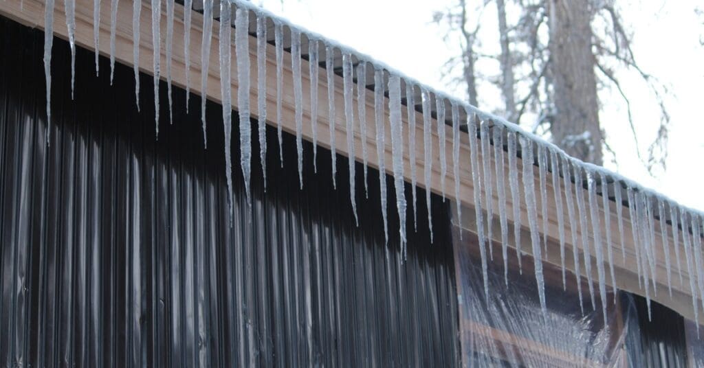 Ice dams form on a light wood roof edge above a dark corrugated wall, with bare trees and an overcast sky in the background.