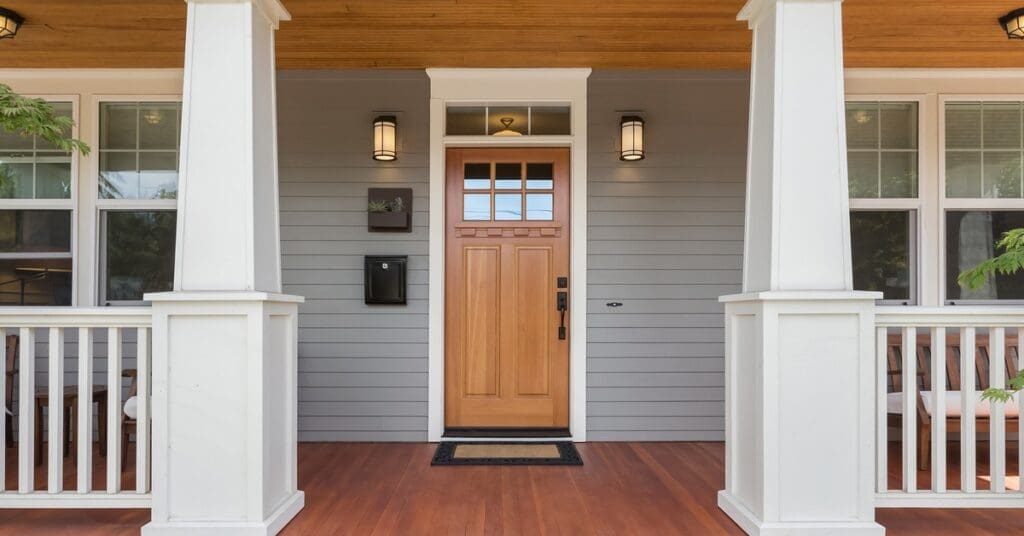 A modern home has a wood porch, brown wood door, white railings, multiple large windows, and light gray vinyl siding.