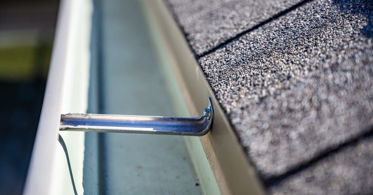 A close-up of a clean white gutter with a metallic hanger below a dark gray asphalt shingle roof, brightly lit by sunlight.