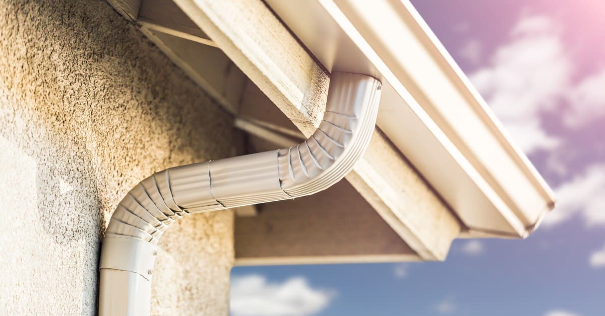 A close-up of a light gray gutter system on a beige stucco wall, with a clear sky and soft sunlight in the background.