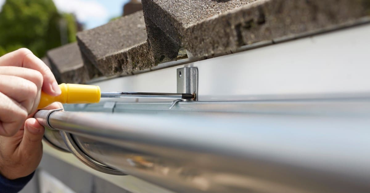 A close-up of hands fixing a silver gutter with a screwdriver under a dark gray roof, with trees in the background.