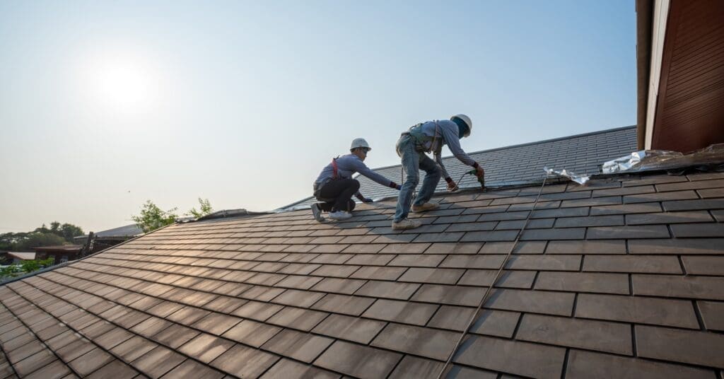 Two roofers wearing safety harnesses install synthetic roofing on a sloped home roof under clear morning sky.