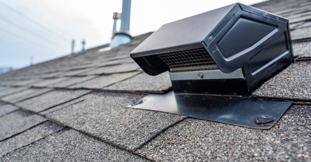 A black roof vent sits on gray asphalt shingles, with metal flashing secured beneath and a chimney in the background.