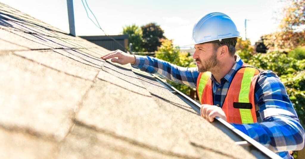 A roofing contractor wearing a hard hat and safety vest inspects the shingles on a home's roof during a bright sunny day.