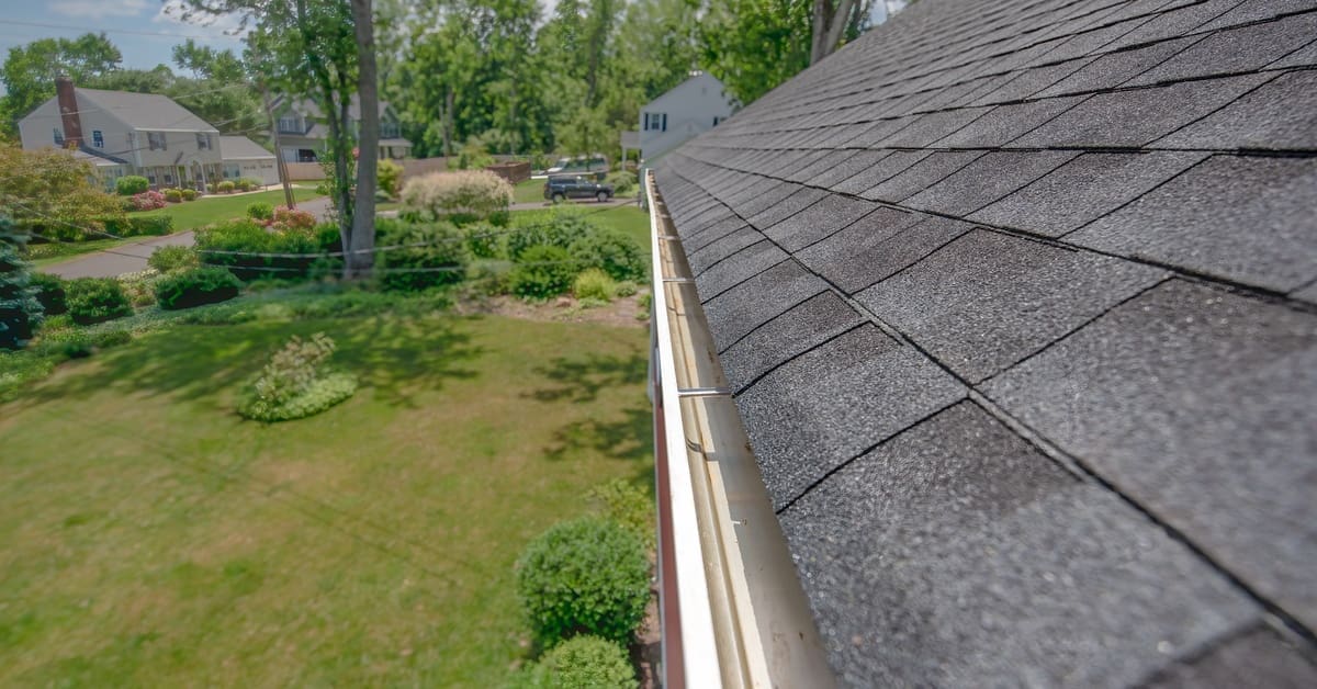 Gray asphalt shingles cover a sloped roof above a white gutter overlooking a green suburban yard with grass and trees.