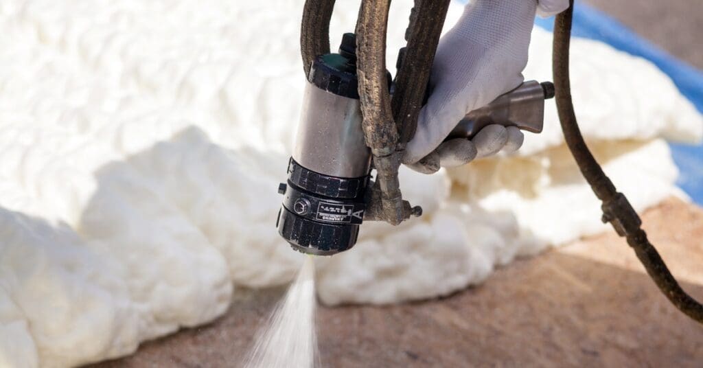 A worker sprays foam insulation from a hose onto a wood surface, forming a thick white barrier along the wall.