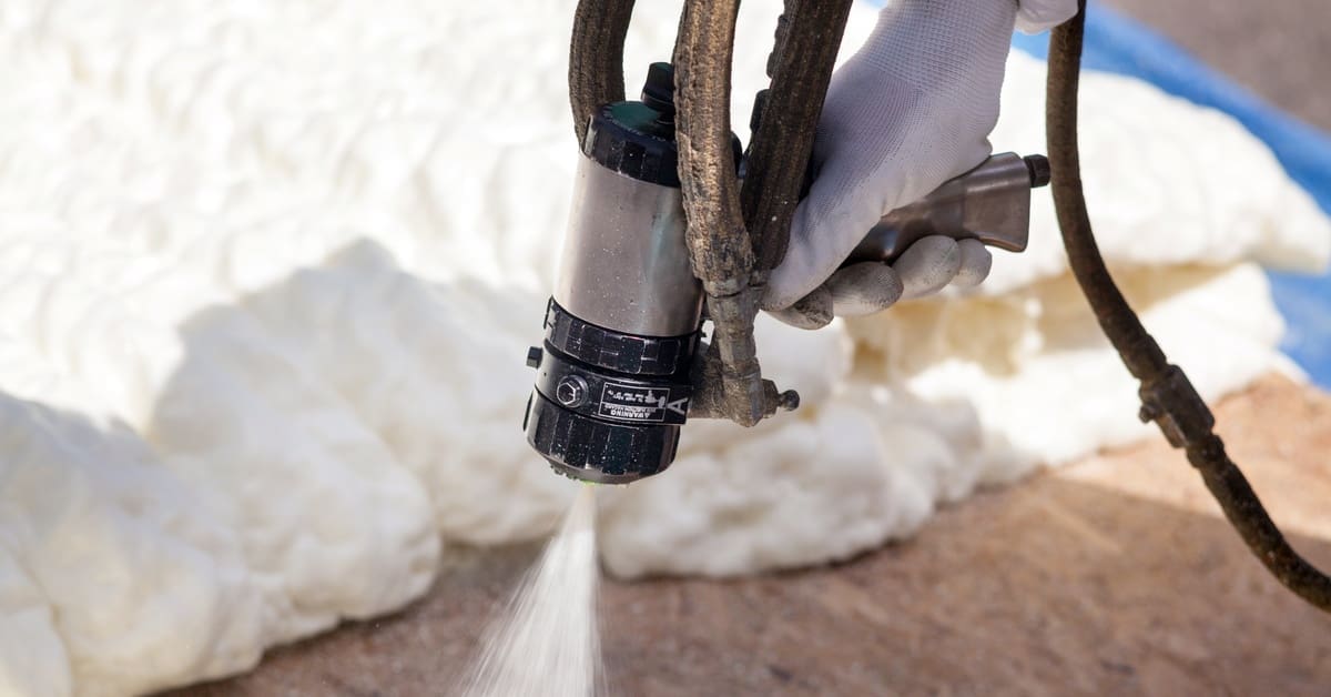 A worker sprays foam insulation from a hose onto a wood surface, forming a thick white barrier along the wall.