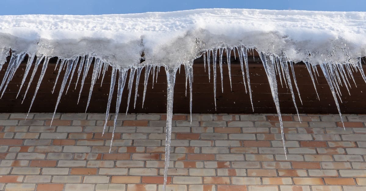 Large icicles hang from a snow-covered roof edge above a brick wall, showing ice dam formation during winter.