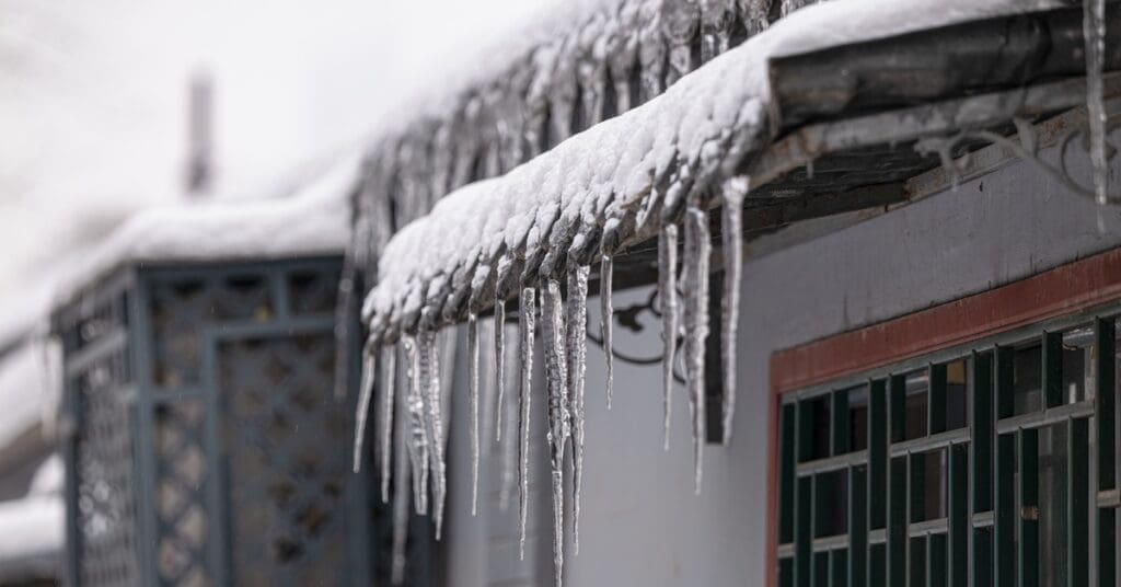 Ice dams hang from a gutter along a snow-covered roof edge, with a window visible on the house's exterior wall.