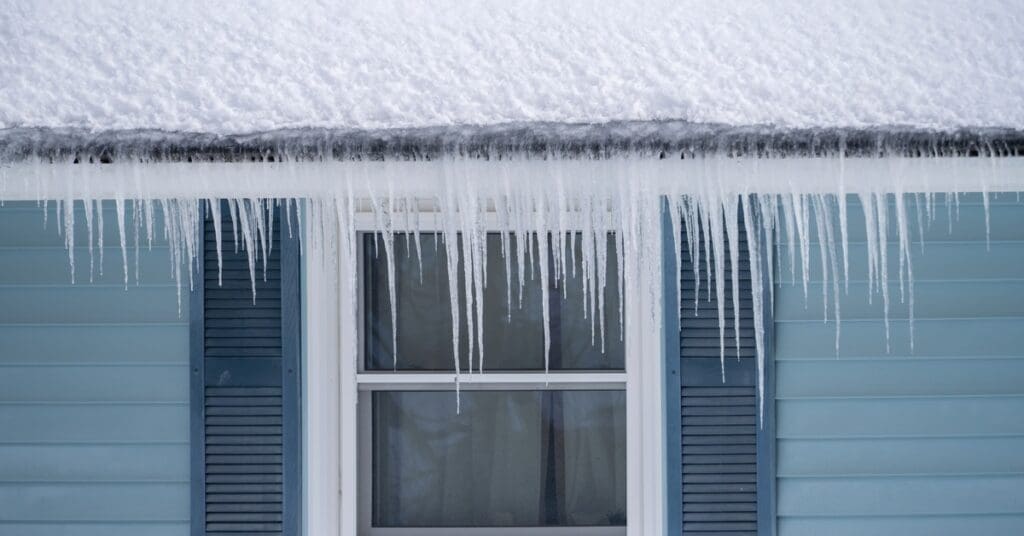 Ice dams hang from a gutter above a window with shutters on a blue house covered in snow during winter.
