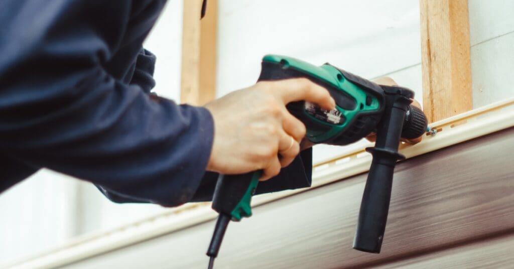 A contractor uses a green and black nail gun to install beige lap siding panels on the exterior of a house frame.