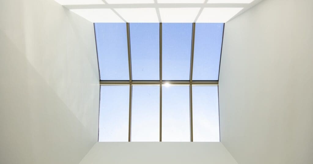 A large rectangular skylight with eight glass panels reveals a clear blue sky through white ceiling beams in a modern room.