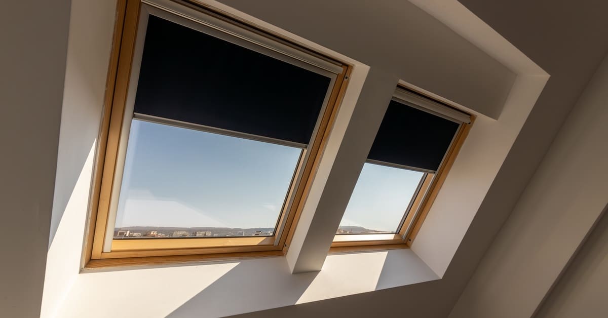 Two wooden-framed skylights with dark roller shades cast angular shadows on white slanted ceiling in a modern attic space.
