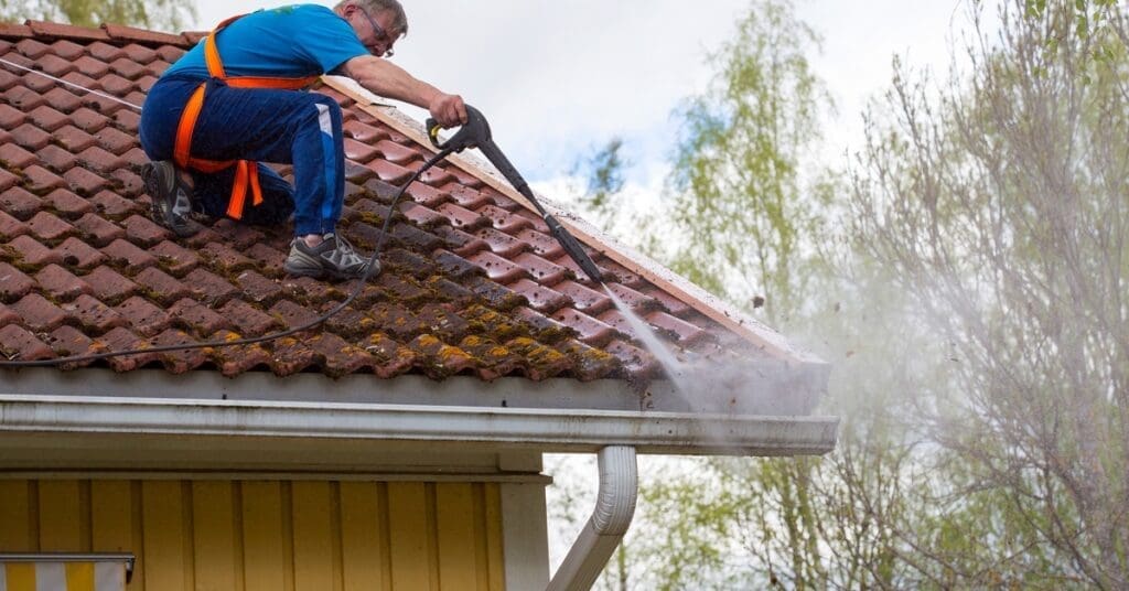 A man wearing safety straps uses a pressure washer to clean debris from a tiled roof and gutter on a home.